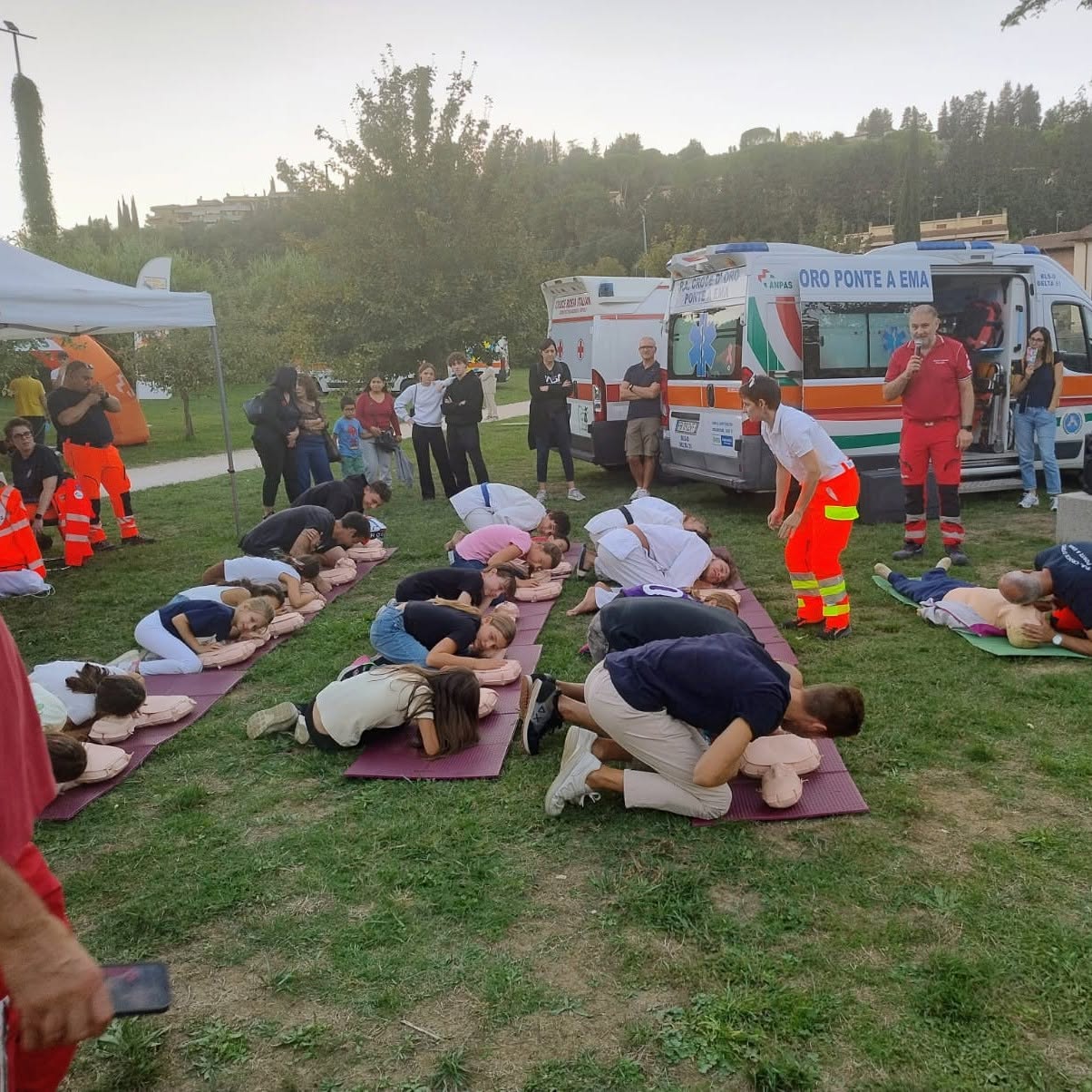 Dimostrazione di rianimazione cardiopolmonare sul prato, con volontari e ambulanze sullo sfondo